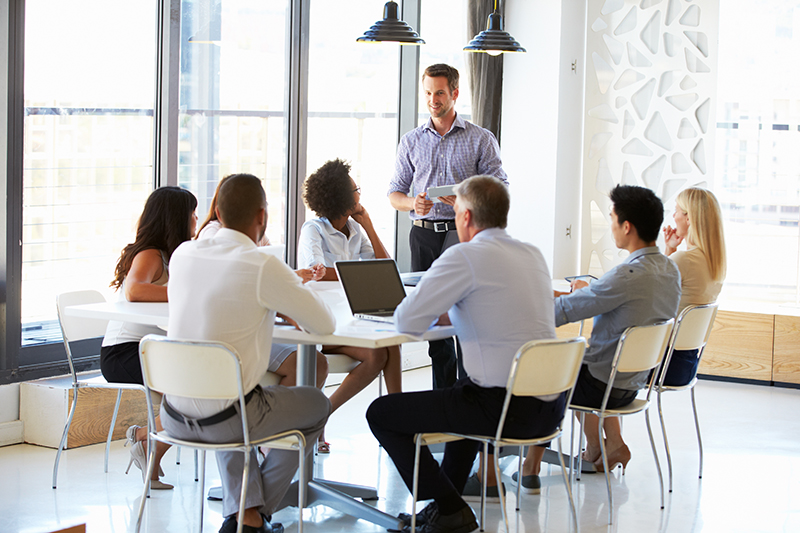 Several people sit at a conference table while a man stands and speaks.