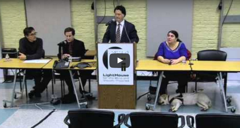 Three people are seated at a table and a man stands at a podium. A guide dog lies under the table.