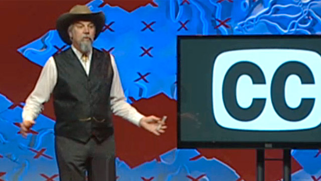 Gary Robson on stage speaking at a TED event. A large tv screen behind him shows the closed caption symbol.