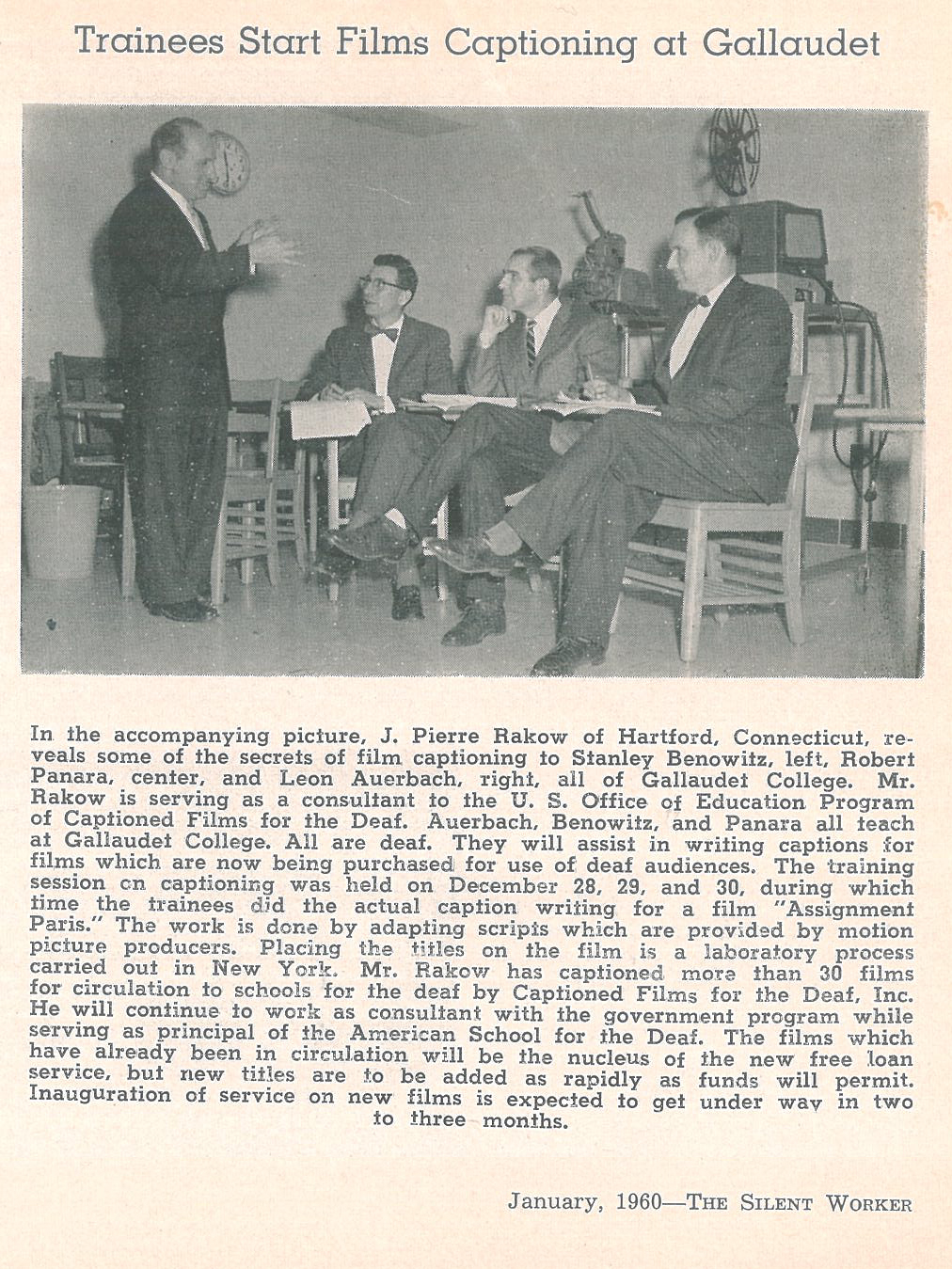 Photo of a man standing talking to three men sitting. A 16 millimeter film projector is behind them.