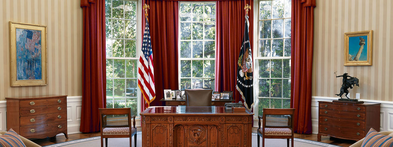 the oval office. a large carved wood desk with american flag behind it and three floor to ceiling windows with heavy drapes.