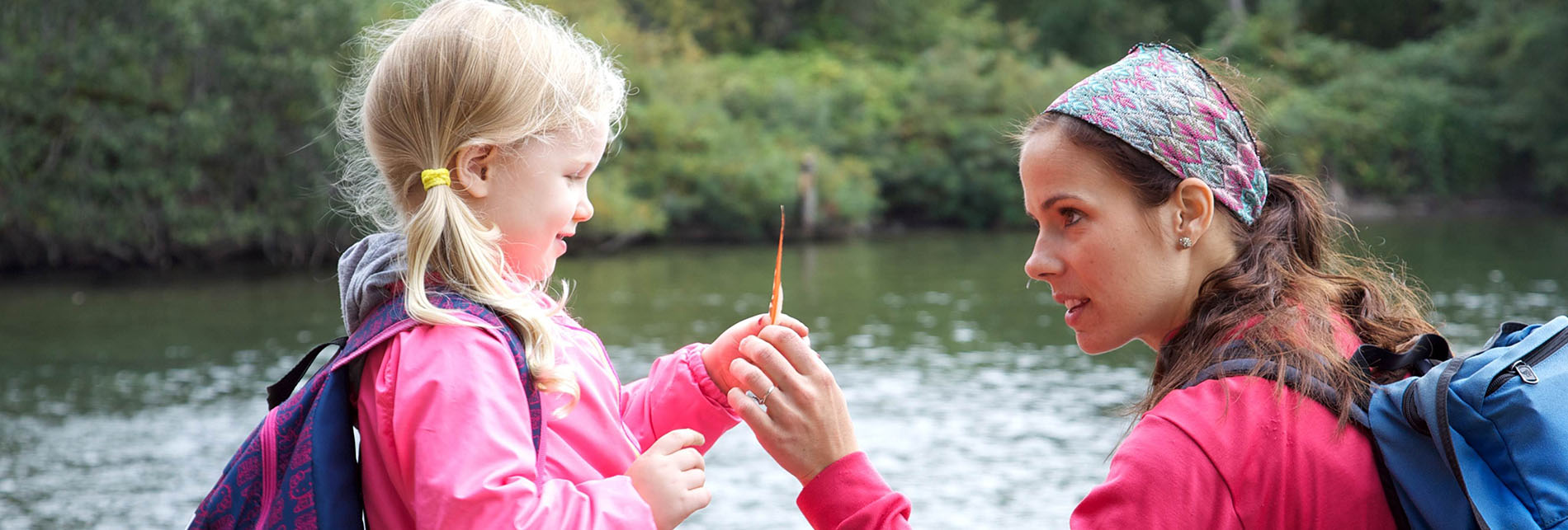 a mother and her young daughter are wearing jackets near a small body of water in a wooded area, looking at an orange leaf or feather held by the daughter.