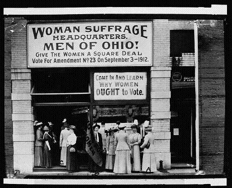 Old Photo of building with large hand-painted sign saying "Woman Suffrage Headquarters. Men of Ohio! Give the women a square deal. Vote for Amendment Number 23 on September 3, 1912. Come in and learn why women OUGHT to vote. Below signs, well-dressed women and men stand outside the entrance.