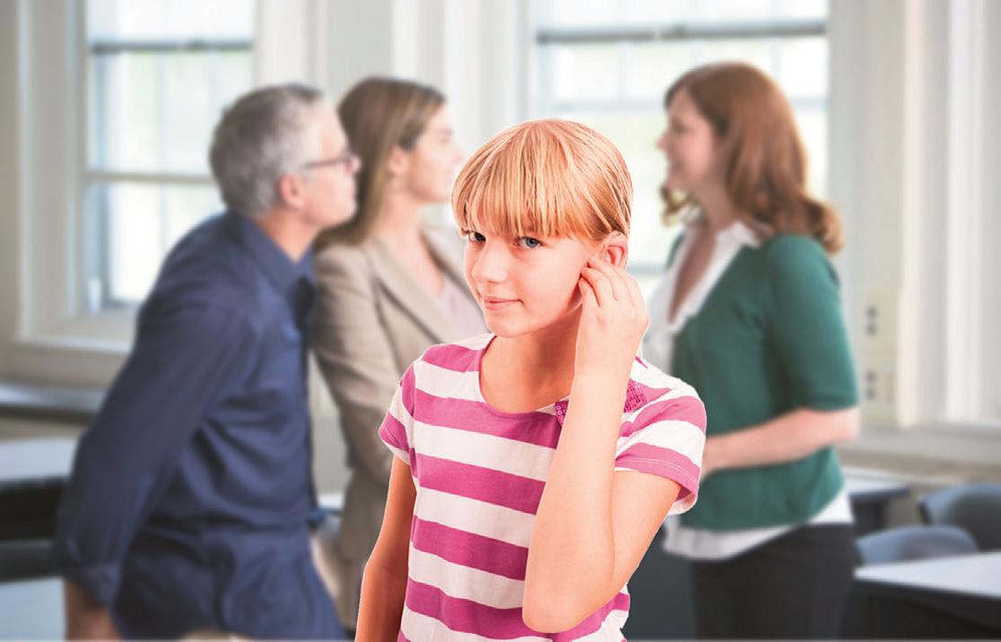 A middle school girl adjusts her hearing aid in a classroom. Behind her, parents and a teacher are communicating.