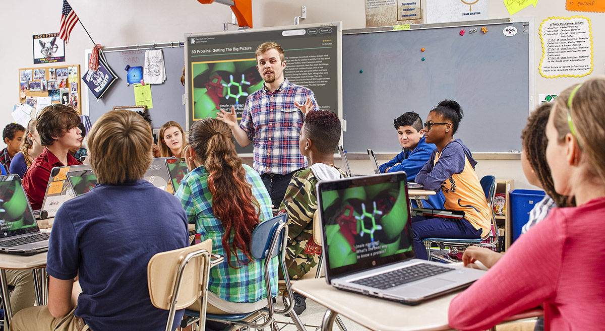 A teacher addresses his students in a classroom. A DCMP video pays on a white board and on students' laptops.