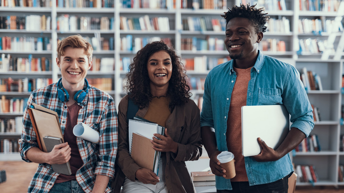 Three young adults in a library and holding books are smiling.