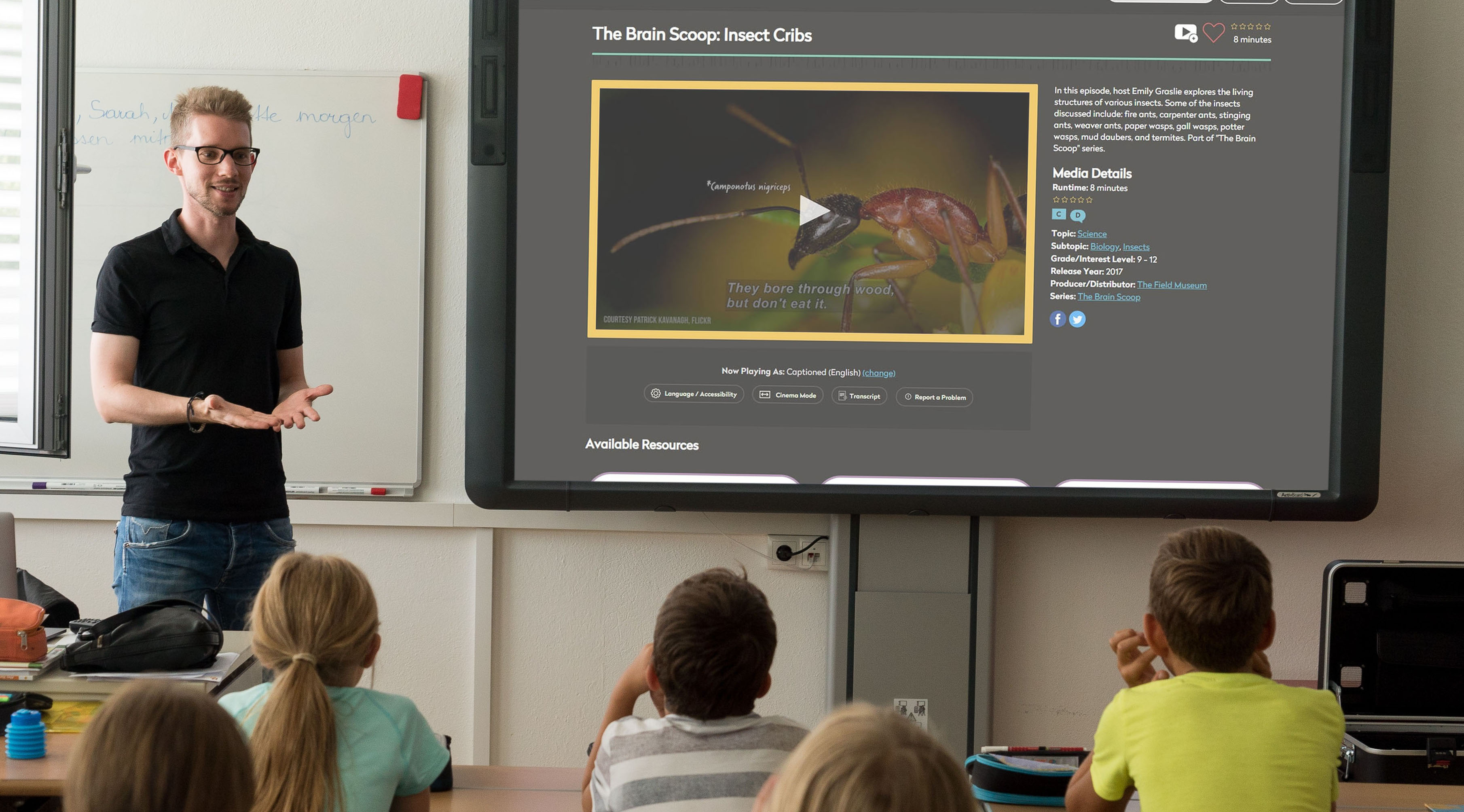 Teacher in a classroom with students. Video is playing on smartboard.