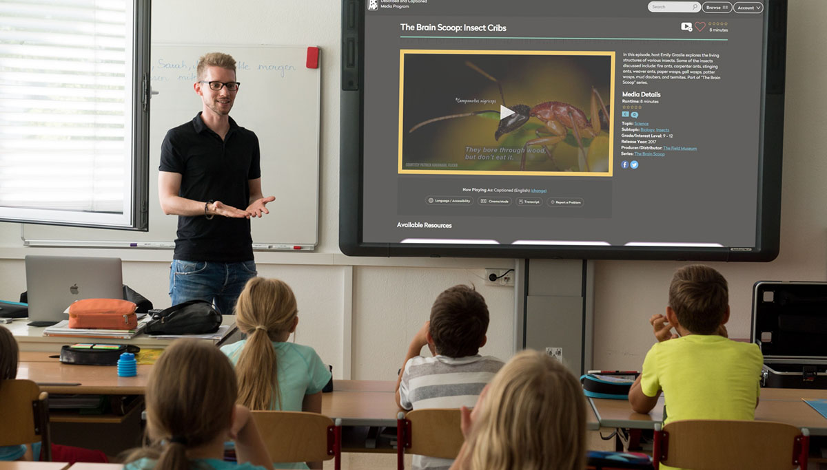 A teacher stands in front of a class of children. A large smartboard on the wall shows the DCMP website with a video.