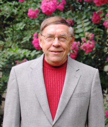 Bill Stark smiles as he poses in front of greeneryi with pink flower. He wears glasses, a red shirt and gray suitcoat.